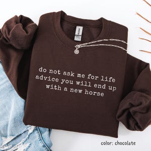 Può includere: Una felpa girocollo color marrone cioccolato con testo bianco che recita: do not ask me for life advice you will end up with a new horse. Il capo include un'etichetta del marchio Gildan all'interno del colletto e una collana d'argento.