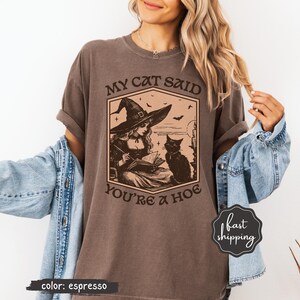 May include: Espresso-colored t-shirt featuring a graphic of a witch reading a book with a black cat. The text on the shirt reads "MY CAT SAID YOU'RE A HOE". The shirt has a vintage, slightly faded look.