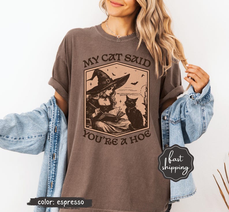 May include: Espresso-colored t-shirt featuring a graphic of a witch reading a book with a black cat. The text on the shirt reads "MY CAT SAID YOU'RE A HOE". The shirt has a vintage, slightly faded look.