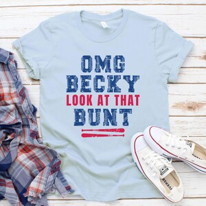Pu&ograve; includere: Una t-shirt a maniche corte color azzurro con la scritta OMG BECKY LOOK AT THAT BUNT in lettere blu e rosse, con una grafica di due mazze da baseball rosse sotto il testo, accompagnata da una camicia di flanella a quadri e scarpe da ginnastica.