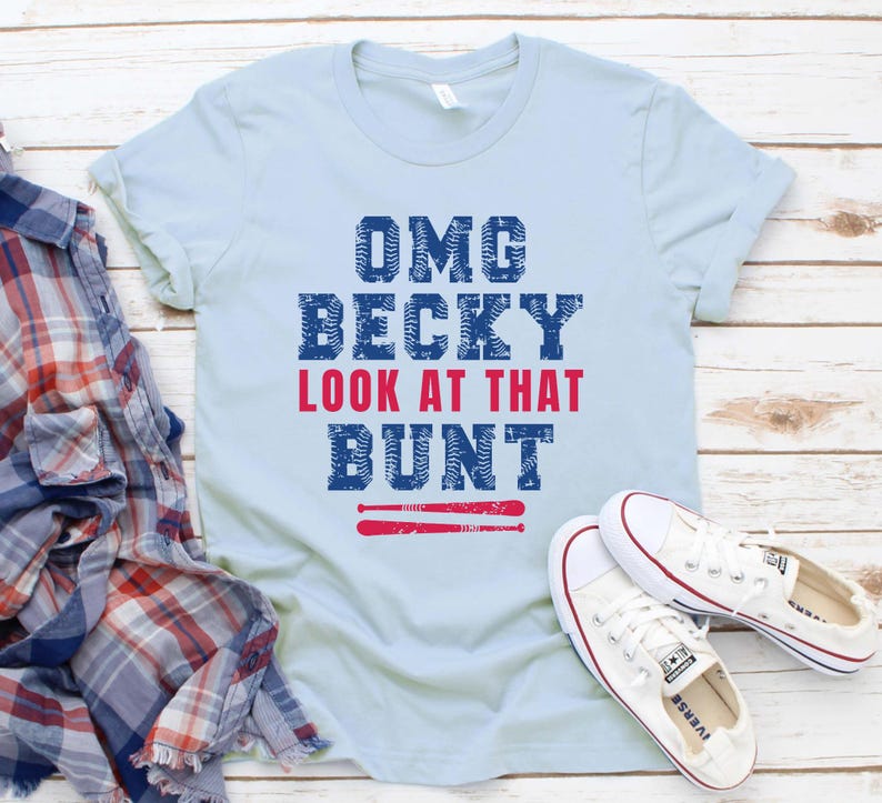 Pu&ograve; includere: Una t-shirt a maniche corte color azzurro con la scritta OMG BECKY LOOK AT THAT BUNT in lettere blu e rosse, con una grafica di due mazze da baseball rosse sotto il testo, accompagnata da una camicia di flanella a quadri e scarpe da ginnastica.