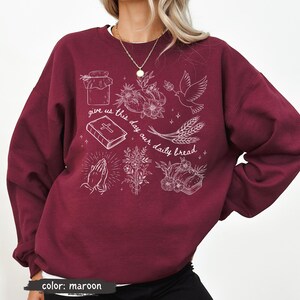 K&ouml;nnte beinhalten: Burgunderfarbenes Sweatshirt mit wei&szlig;en grafischen Illustrationen, darunter ein Glas, Blumen, eine Taube, Weizen, ein Buch, betende H&auml;nde und der Text "give us this day our daily bread."