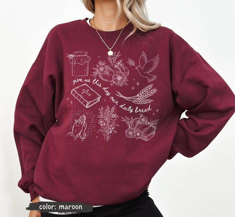K&ouml;nnte beinhalten: Burgunderfarbenes Sweatshirt mit wei&szlig;en grafischen Illustrationen, darunter ein Glas, Blumen, eine Taube, Weizen, ein Buch, betende H&auml;nde und der Text "give us this day our daily bread."