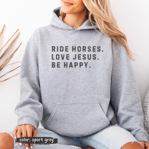 May include: A light gray hoodie with a front pocket and a drawstring hood. The text "RIDE HORSES. LOVE JESUS. BE HAPPY." is printed in a distressed black font. The bottom left corner of the image says "color: sport grey."