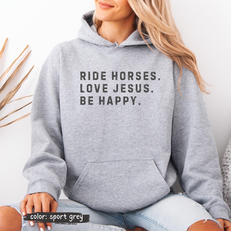 May include: A light gray hoodie with a front pocket and a drawstring hood. The text "RIDE HORSES. LOVE JESUS. BE HAPPY." is printed in a distressed black font. The bottom left corner of the image says "color: sport grey."