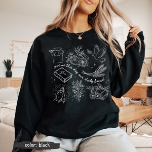 K&ouml;nnte beinhalten: Schwarzes Sweatshirt mit wei&szlig;en grafischen Illustrationen eines Glases, Blumen, einer Taube, Weizen, eines Buches, betenden H&auml;nden und Brot. Der Text "give us this day our daily bread" ist ebenfalls enthalten.