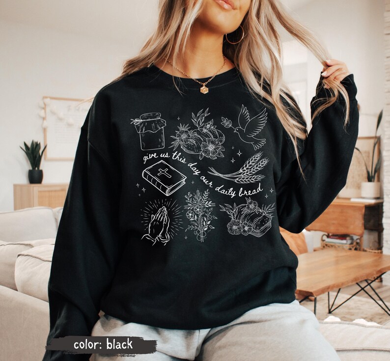 K&ouml;nnte beinhalten: Schwarzes Sweatshirt mit wei&szlig;en grafischen Illustrationen eines Glases, Blumen, einer Taube, Weizen, eines Buches, betenden H&auml;nden und Brot. Der Text "give us this day our daily bread" ist ebenfalls enthalten.