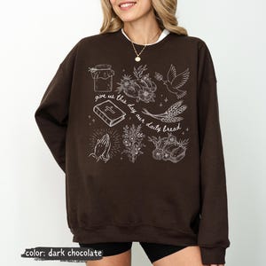 K&ouml;nnte beinhalten: Dunkel schokoladenfarbenes Sweatshirt mit wei&szlig;en grafischen Illustrationen. Das Design umfasst ein Glas, Blumen, eine Taube, Weizen, ein Buch, betende H&auml;nde und den Text "give us this day our daily bread."