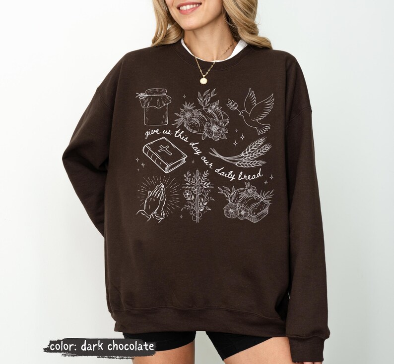 K&ouml;nnte beinhalten: Dunkel schokoladenfarbenes Sweatshirt mit wei&szlig;en grafischen Illustrationen. Das Design umfasst ein Glas, Blumen, eine Taube, Weizen, ein Buch, betende H&auml;nde und den Text "give us this day our daily bread."