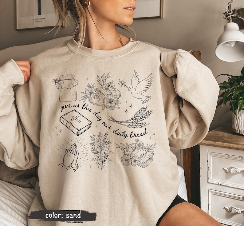 May include: Sand-colored sweatshirt with a black graphic design featuring a jar, flowers, a dove, wheat, a book, praying hands, and bread. The text "give us this day our daily bread" is also included. The sweatshirt is a neutral color.