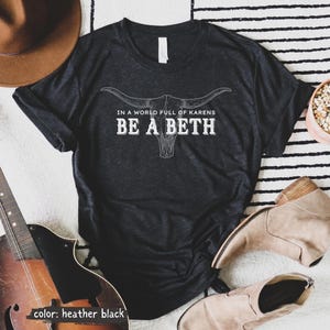 May include: A heather black short-sleeve t-shirt featuring a white graphic of a longhorn skull. The text printed in white reads, "IN A WORLD FULL OF KARENS BE A BETH." The shirt has a crew neckline and is displayed with a brown hat and tan boots.