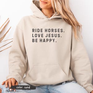 May include: Sand-colored hoodie with a front pocket and drawstring hood. The text "RIDE HORSES. LOVE JESUS. BE HAPPY." is printed in a dark gray font. The hoodie is a neutral color and suitable for casual wear.