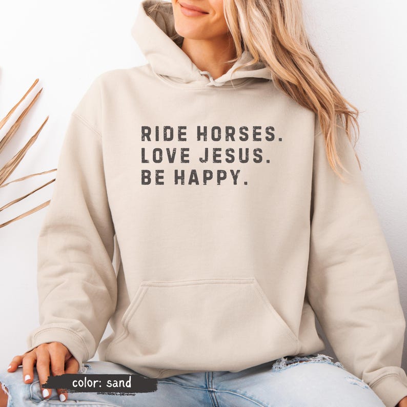 May include: Sand-colored hoodie with a front pocket and drawstring hood. The text "RIDE HORSES. LOVE JESUS. BE HAPPY." is printed in a dark gray font. The hoodie is a neutral color and suitable for casual wear.