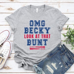 Pu&ograve; includere: Una t-shirt grigio m&eacute;lange a maniche corte con la scritta blu e rossa "OMG BECKY LOOK AT THAT BUNT" e un grafico di due mazze da baseball sottostanti. Le lettere presentano un motivo testurizzato che ricorda le cuciture di una palla.