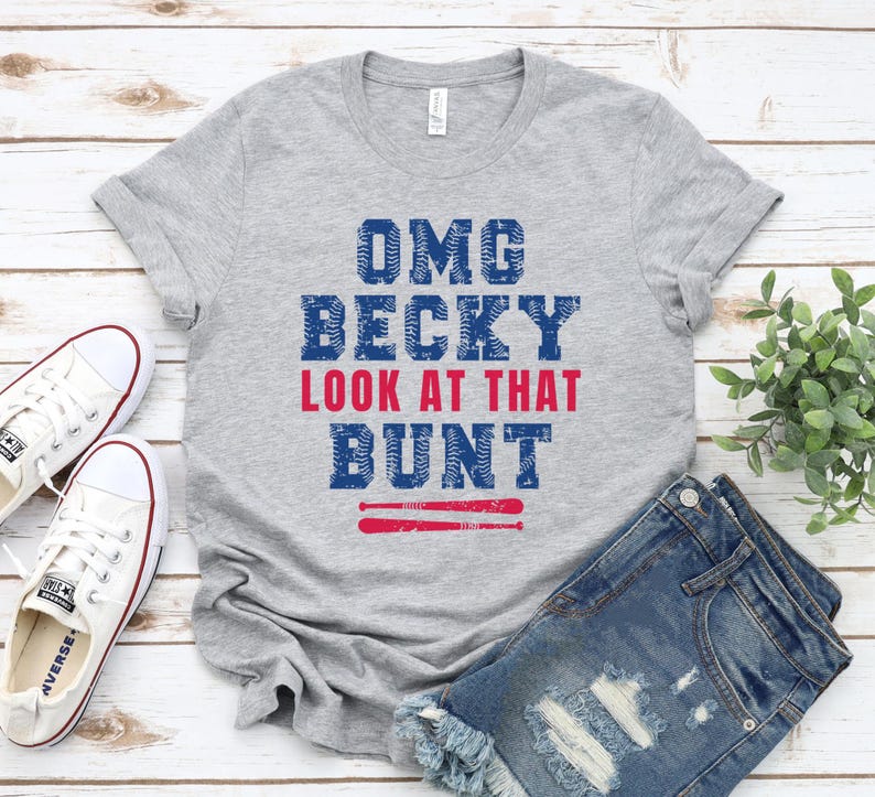 Pu&ograve; includere: Una t-shirt grigio m&eacute;lange a maniche corte con la scritta blu e rossa "OMG BECKY LOOK AT THAT BUNT" e un grafico di due mazze da baseball sottostanti. Le lettere presentano un motivo testurizzato che ricorda le cuciture di una palla.