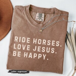 May include: Espresso-colored t-shirt with the text "RIDE HORSES. LOVE JESUS. BE HAPPY." A silver necklace with a small pendant rests on the shirt. The shirt is a Comfort Colors brand, size S-P-CH.