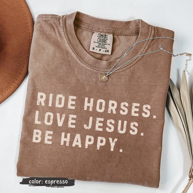 May include: Espresso-colored t-shirt with the text "RIDE HORSES. LOVE JESUS. BE HAPPY." A silver necklace with a small pendant rests on the shirt. The shirt is a Comfort Colors brand, size S-P-CH.