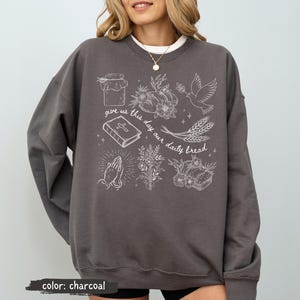 K&ouml;nnte beinhalten: Anthrazitfarbenes Sweatshirt mit wei&szlig;en grafischen Illustrationen eines Glases, Blumen, einer Taube, Weizen, eines Buches, betenden H&auml;nden und Brot. Der Text "give us this day our daily bread" ist ebenfalls aufgedruckt.
