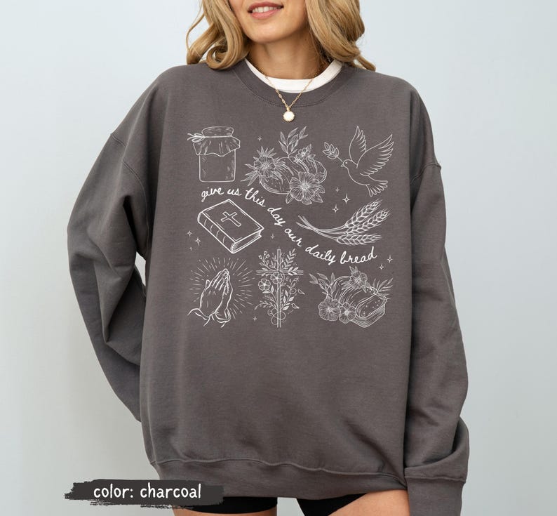 K&ouml;nnte beinhalten: Anthrazitfarbenes Sweatshirt mit wei&szlig;en grafischen Illustrationen eines Glases, Blumen, einer Taube, Weizen, eines Buches, betenden H&auml;nden und Brot. Der Text "give us this day our daily bread" ist ebenfalls aufgedruckt.