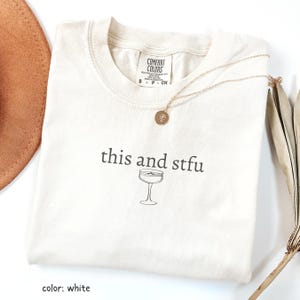 Może przedstawiać: Biały t-shirt z napisem "this and stfu" i grafiką kieliszka do koktajli. Złoty naszyjnik z wisiorkiem leży na koszulce. Koszulka ma metkę Comfort Colors. Brązowy kapelusz i suszona roślina są w tle.