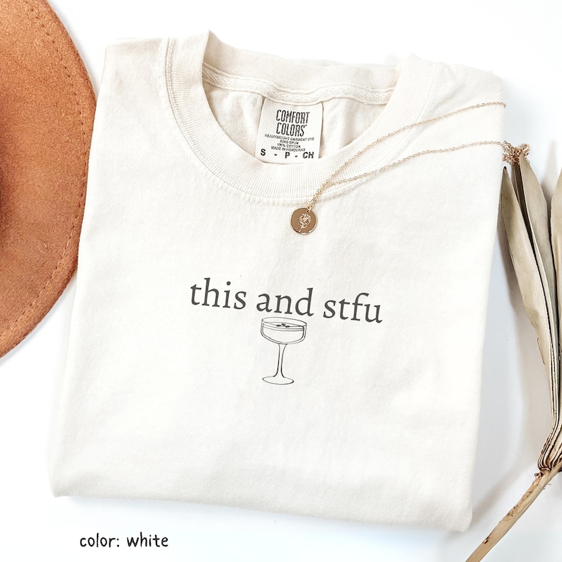 Może przedstawiać: Biały t-shirt z napisem "this and stfu" i grafiką kieliszka do koktajli. Złoty naszyjnik z wisiorkiem leży na koszulce. Koszulka ma metkę Comfort Colors. Brązowy kapelusz i suszona roślina są w tle.