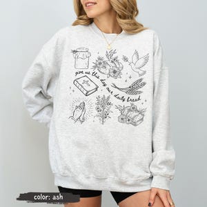 K&ouml;nnte beinhalten: Hellgraues Sweatshirt mit Rundhalsausschnitt und langen &Auml;rmeln. Auf der Vorderseite befinden sich schwarze Strichzeichnungen von einem Glas, Blumen, einer Taube, einer Bibel, betenden H&auml;nden, Weizen und Brot. Der Text "give us this day our daily bread" ist ebenfalls enthalten.