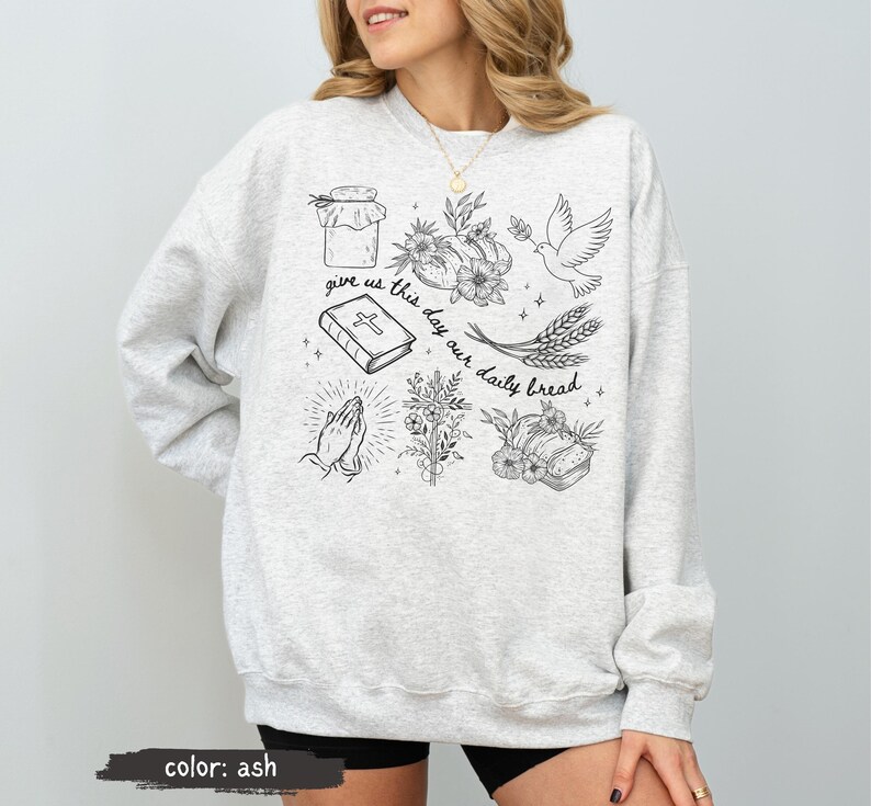 K&ouml;nnte beinhalten: Hellgraues Sweatshirt mit Rundhalsausschnitt und langen &Auml;rmeln. Auf der Vorderseite befinden sich schwarze Strichzeichnungen von einem Glas, Blumen, einer Taube, einer Bibel, betenden H&auml;nden, Weizen und Brot. Der Text "give us this day our daily bread" ist ebenfalls enthalten.