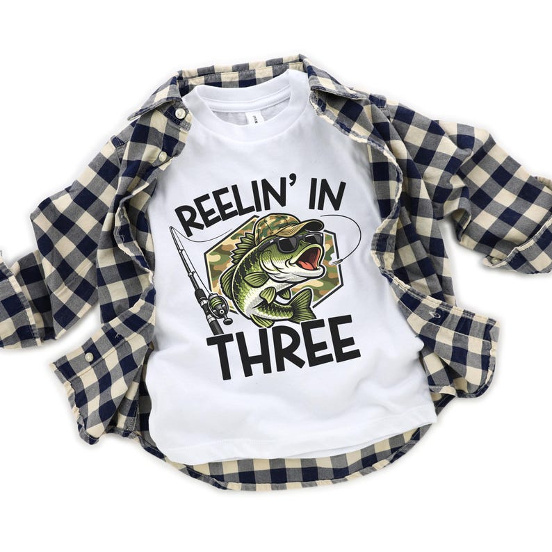 Puede incluir: Camiseta blanca con un gr&aacute;fico de un pez con gafas de sol y un sombrero de camuflaje, con el texto "Reelin' In Three". La camiseta est&aacute; superpuesta con una camisa abotonada a cuadros azules y blancos.