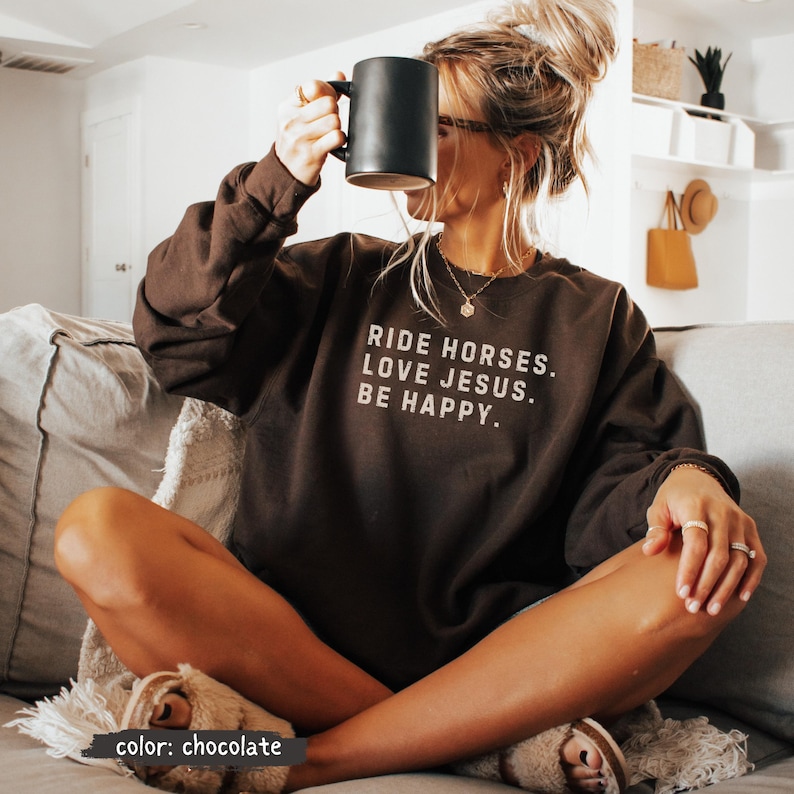 May include: Chocolate-colored sweatshirt with the text "RIDE HORSES. LOVE JESUS. BE HAPPY." in white. The sweatshirt is worn with a black mug. The color of the sweatshirt is labeled "chocolate".