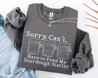 May include: Charcoal gray sweatshirt with the text "Sorry. Can't. Have to Feed My Sourdough Starter" in white. The design includes three jars with sourdough starter. A silver necklace with a pendant is draped across the sweatshirt. The color of the sweatshirt is charcoal.