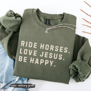 May include: Military green sweatshirt with the text "RIDE HORSES. LOVE JESUS. BE HAPPY." A silver necklace with a circular pendant is draped across the sweatshirt. The sweatshirt is folded on top of a pair of blue jeans.
