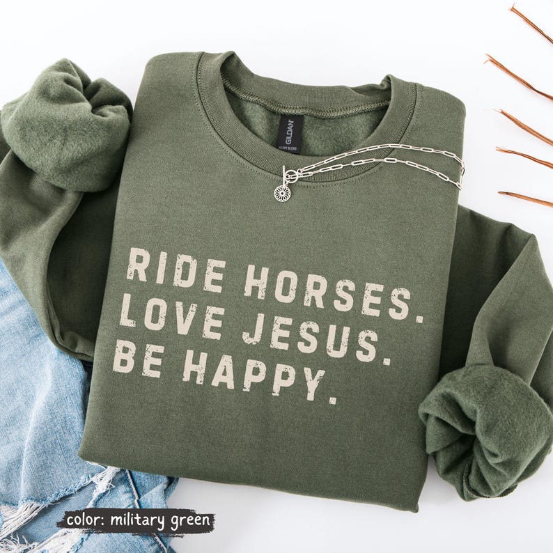 May include: Military green sweatshirt with the text "RIDE HORSES. LOVE JESUS. BE HAPPY." A silver necklace with a circular pendant is draped across the sweatshirt. The sweatshirt is folded on top of a pair of blue jeans.