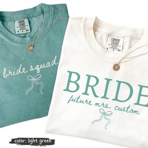 Può includere: Una t-shirt verde acqua con testo bianco bride squad e un grafico a fiocco, e una t-shirt bianca con testo verde acqua BRIDE future mrs. custom e un grafico a fiocco. Entrambe le maglie hanno un'etichetta Comfort Colors e una collana dorata.