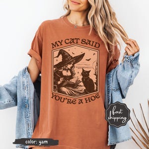 May include: A yam-colored t-shirt featuring a graphic of a witch in a hat reading a book with a black cat. The text on the shirt reads "MY CAT SAID YOU'RE A HOE". The shirt has rolled-up sleeves.
