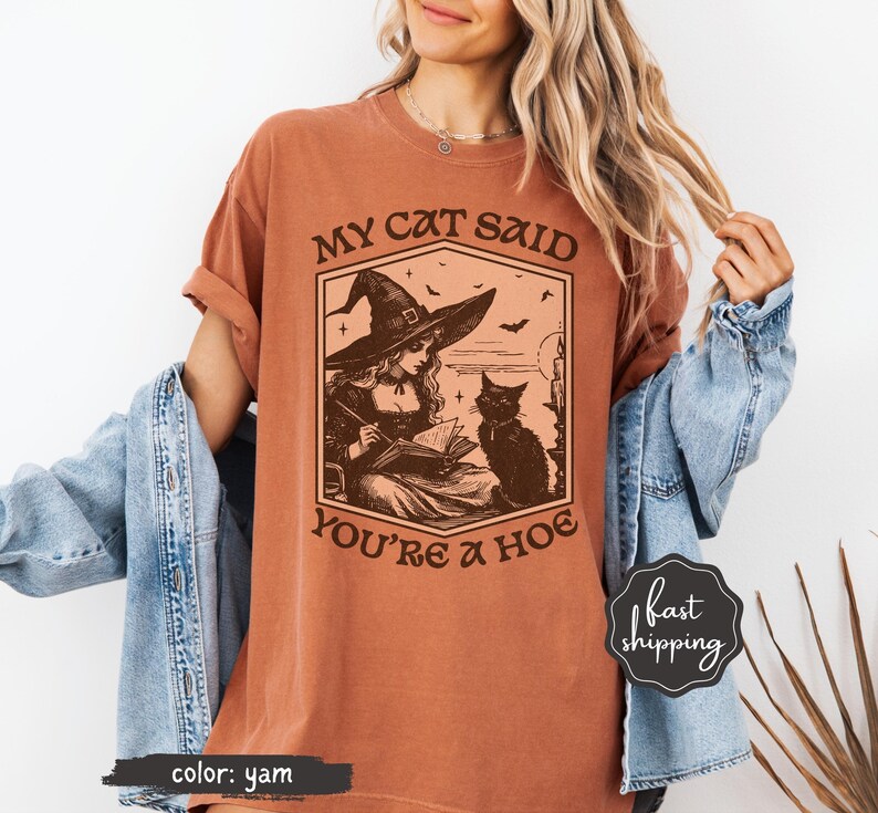 May include: A yam-colored t-shirt featuring a graphic of a witch in a hat reading a book with a black cat. The text on the shirt reads "MY CAT SAID YOU'RE A HOE". The shirt has rolled-up sleeves.