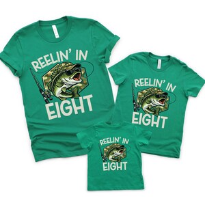 Puede incluir: Tres camisetas verdes con el texto "REELIN' IN EIGHT" y un gr&aacute;fico de un pez con gafas de sol y un sombrero de camuflaje. Las camisetas son de diferentes tallas, lo que sugiere un conjunto familiar.