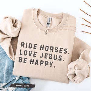 May include: Sand-colored sweatshirt with the text "RIDE HORSES. LOVE JESUS. BE HAPPY." in dark gray. A silver necklace with a small pendant rests on the shirt. The sweatshirt is folded on top of a pair of blue jeans and a beige fabric.