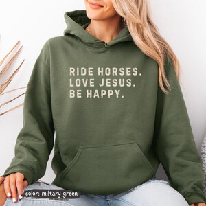May include: Military green hoodie with the text "RIDE HORSES. LOVE JESUS. BE HAPPY." in a white, sans-serif font. The hoodie has a kangaroo pocket and a drawstring hood. The color is described as "military green".
