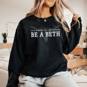 May include: Black sweatshirt with a white graphic design that says "In a world full of Karens, Be a Beth" with a stylized cow skull graphic.