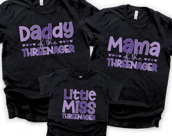 May include: Three black short-sleeve t-shirts with purple graphic text. The shirts read Daddy of the Threenager, Mama of the Threenager, and Little Miss Threenager, each accented with small purple heart icons between the lines of text.