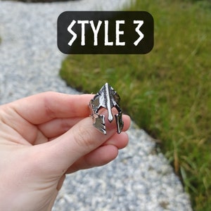 Pu&ograve; includere: Un anello d'argento con un designo a forma di elmo spartano. L'anello &egrave; indossato su un dito, con uno sfondo sfocato di un ruscello e erba verde. Il testo "STYLE 3" &egrave; visualizzato nella parte superiore centrale dell'immagine.