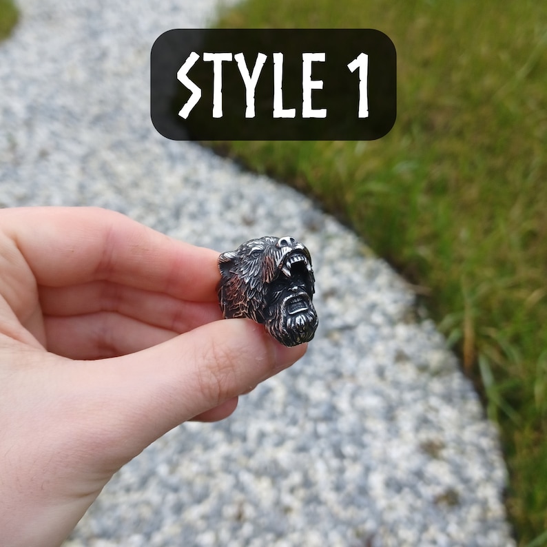 Pu&ograve; includere: Un anello d'argento con un designo dettagliato di una testa d'orso con la bocca aperta e la barba. L'anello &egrave; tenuto in una mano su uno sfondo di erba verde e un sentiero di pietra.