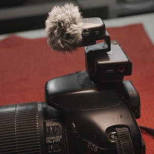 May include: A black DSLR camera with a lens and a microphone attached. The microphone has a fluffy, grey windscreen. The camera is on a red surface.