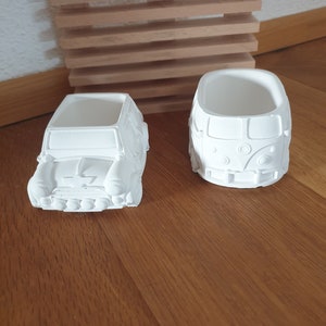 Może przedstawiać: Dwa białe ceramiczne doniczki w kształcie zabytkowych samochodów. Doniczka po lewej stronie ma kształt Mini Coopera, a doniczka po prawej stronie ma kształt furgonetki Volkswagena.