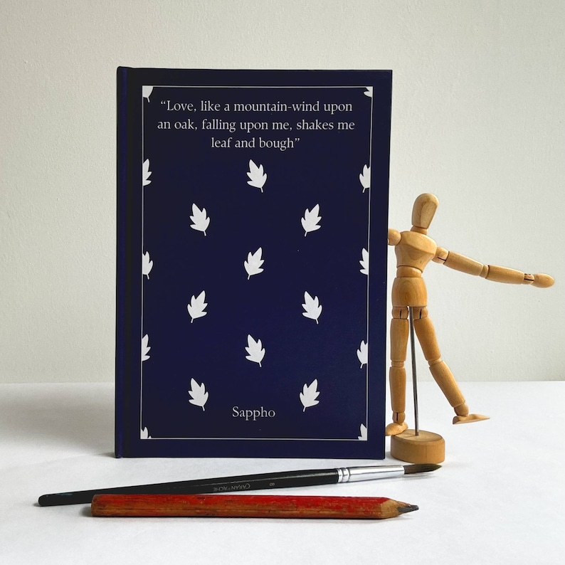 May include: A navy blue notebook with white leaf designs and the text "Love, like a mountain-wind upon an oak, falling upon me, shakes me leaf and bough" and "Sappho" printed on the cover.