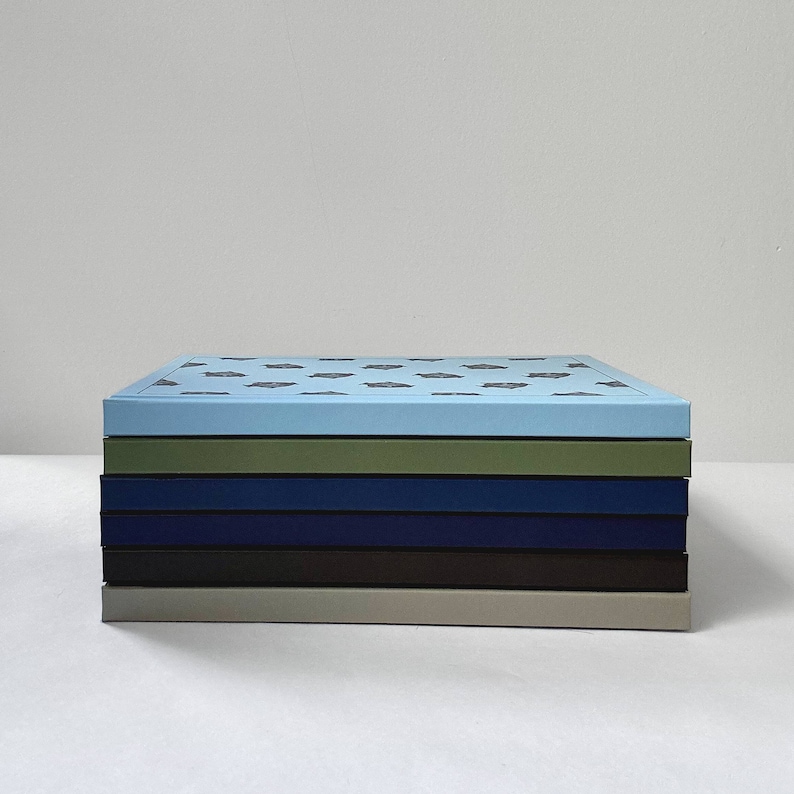 May include: A stack of five notebooks with different colored covers. The top notebook has a light blue cover with a pattern of small, dark blue shapes. The other notebooks have green, dark blue, brown, and light gray covers.