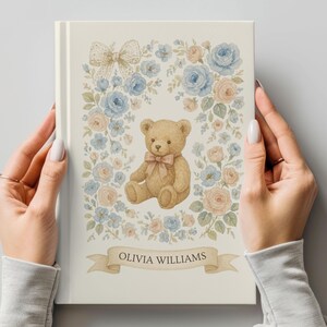 May include: A cream-colored book cover featuring a watercolor illustration of a teddy bear with a bow, surrounded by blue and peach roses. A banner at the bottom reads "OLIVIA WILLIAMS". The design includes a bow at the top. The book is held by two hands.
