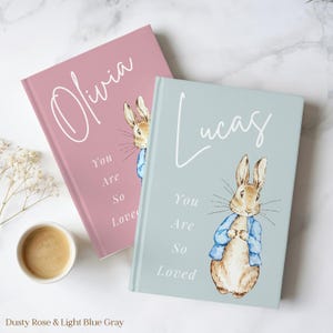 May include: Two personalized baby journals, one dusty rose and one light blue gray. Each journal features a watercolor illustration of a rabbit in a blue jacket. The names "Olivia" and "Lucas" are written in cursive, with the phrase "You Are So Loved" below.