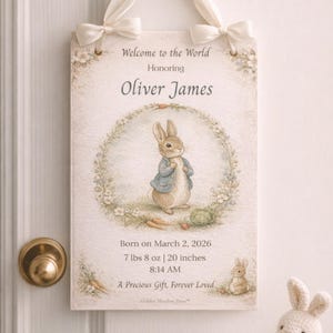 May include: A rectangular sign with a cream background, featuring a watercolor illustration of a rabbit in a blue jacket. The sign includes the text "Welcome to the World Honoring Oliver James." It also states the baby's birth date, weight (3.57 kg), and height (50.8 cm).