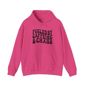 Könnte beinhalten: Ein rosa Hoodie mit einem schwarzen Aufdruck, der in einer Retro-Schrift "Fueled by Caffeine & Chaos" sagt.
